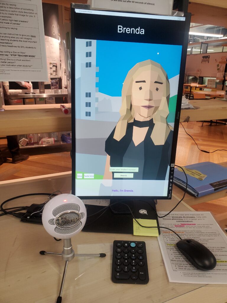 Picture 1 - Our AI conversation partner setup - a screen with the avatar of a character named Brenda. In front of the screen is a mouse, a small keyboard, and a microphone.