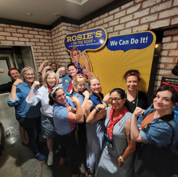 Picture 2 - IALLT members as Rosie the Riveter - women wearing work shirts and flexing their arms in front of a sign that says "We Can Do It!"Photo credit: Lauren Rosen